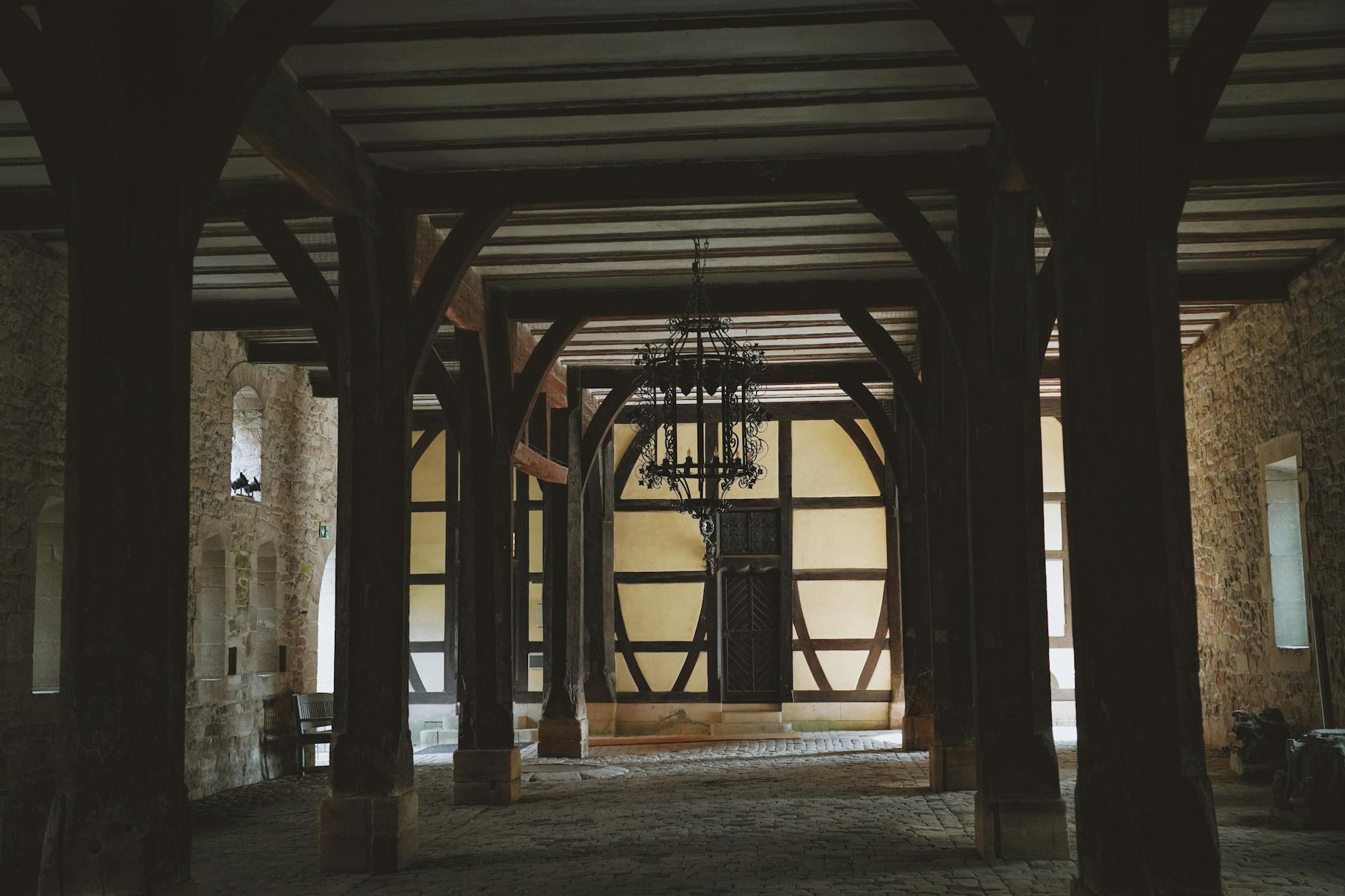 Within Stone and Shadow The Enduring Spell of Castle Interiors 3 - Grand Boudoir A rustic interior with timber beams, stone walls, and an ornate chandelier in an abandoned hall.