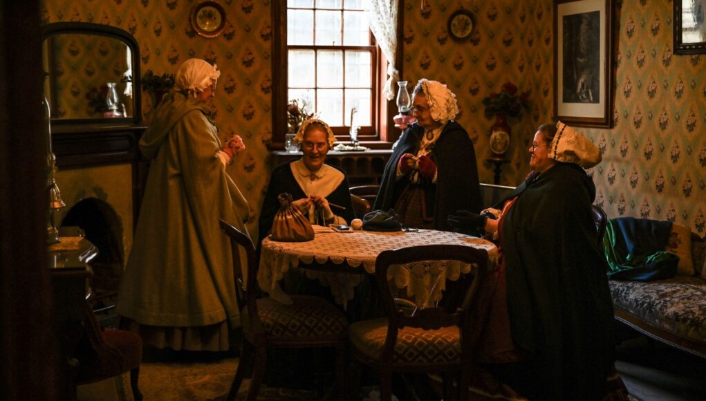 Victorian women in a cozy parlor engaged in conversation, dressed in period attire.