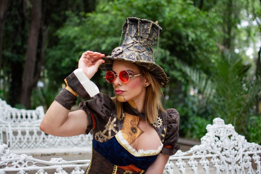 Woman in steampunk costume with hat and glasses in a lush outdoor setting.