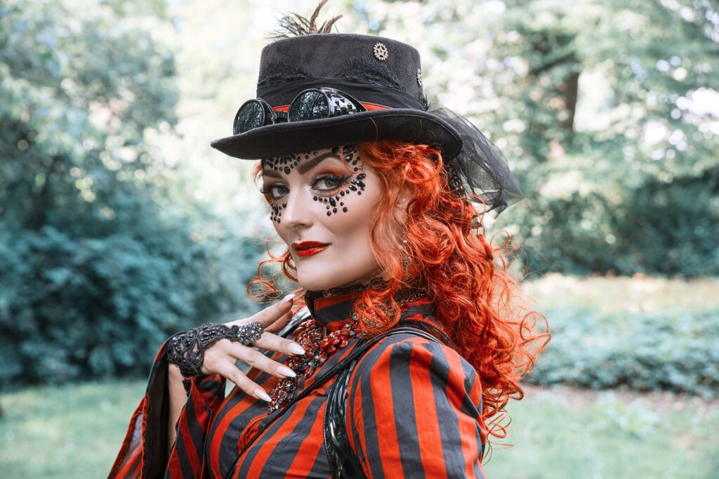 A vibrant steampunk cosplay with intricate face art and orange hair, set outdoors in summer.