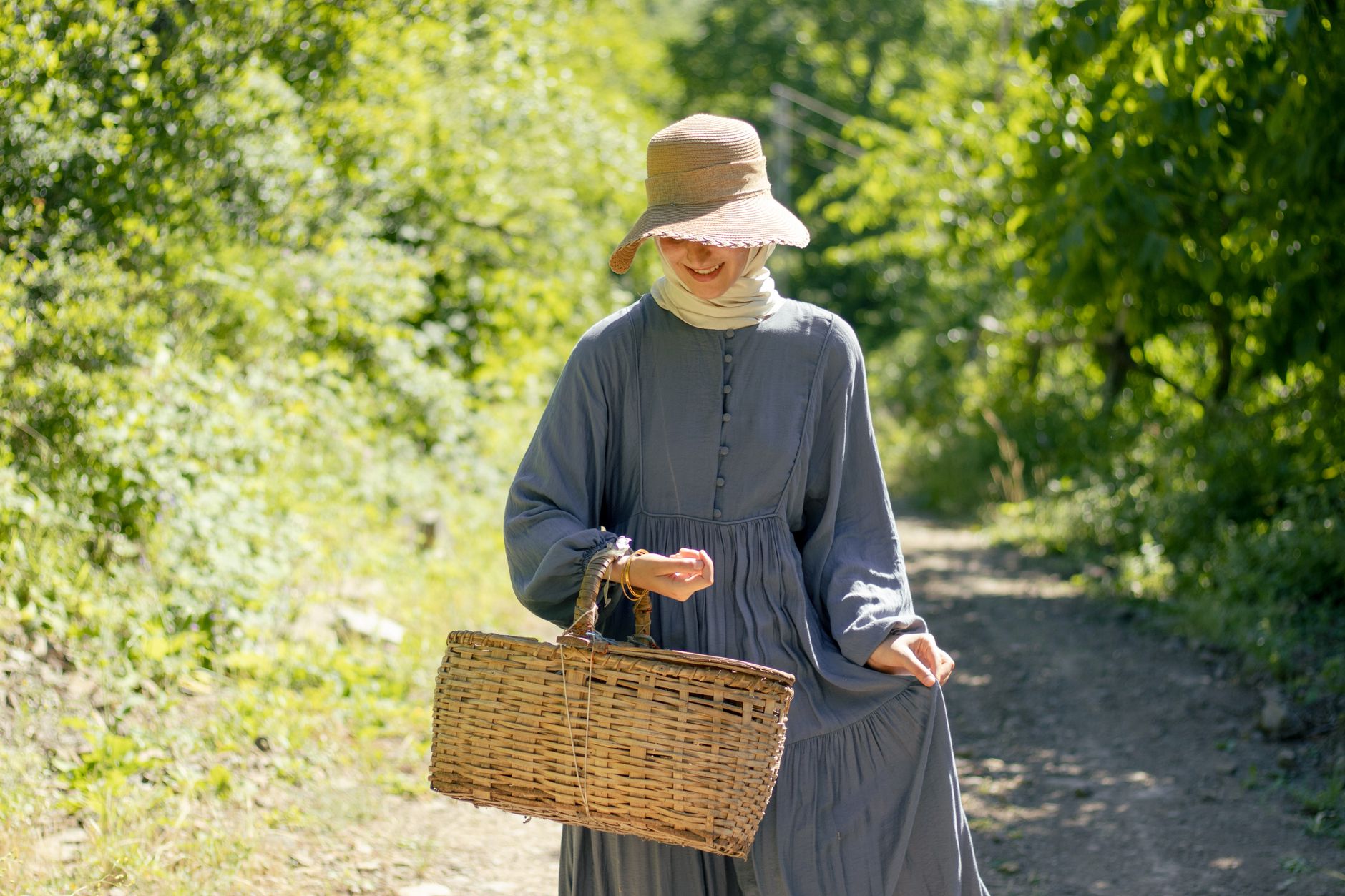 pexels photo 26830925 - A woman in a sun hat walks on a sunny path with a woven basket, surrounded by lush greenery.